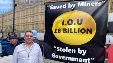 Campaigner Mick Newton outside the Houses of Parliament with a banner which reads " Saved by Miners, I.O.U £8 billion, Stolen by Government".