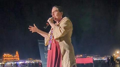 Lisa is shown holding a microphone and speaking during the event. It is dark, and the city lights are visible in the background. She is wearing a long red dress and a cream jacket, and has one hand up as she is talking. 