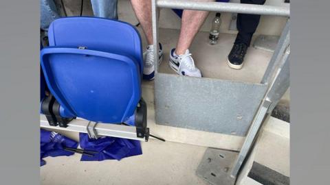 A row of seats at Everton's Hill Dickinson stadium shows a metal panel and railing where a seat should have been. It is at the end of a row of blue fold-up plastic seats.
