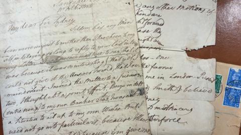 The two letters with handwritten text on folded white paper on top of a manilla envelope with blue and green postage stamps. The letter on the left has several ink stains. The letter on the right is partially damaged with a wide tear halfway down. The handwriting is in neat lines and written with a flourish.