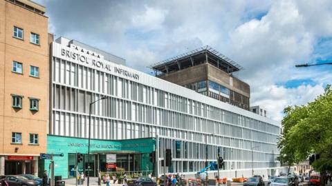 The Image shows the hospital building from the outside, the building is located next to a busy road with cars on it. The outside of the building is turquoise. 