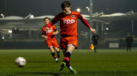 An FC IOM player kicks the ball while another player runs along behind him watching it.
