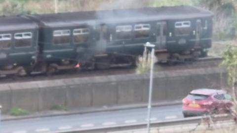 A train carriage has a fire underneath it with smoke emanating. In front of the train line is a road with one car driving past. The photo is hazy as if taken through a window.