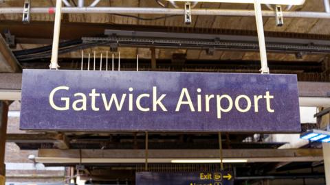 Sign at Railway station with platform and leaving white train of Thames Link at Gatwick Airport London on a cloudy summer day.