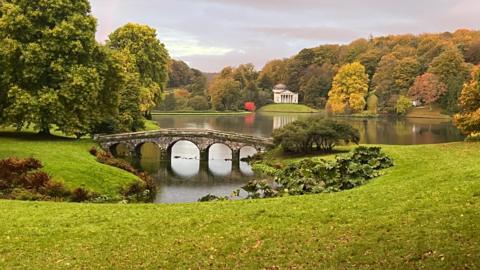 Part of Stourhead's landscape gardens on a cloudy day. Green lawn in front of a lake surrounded by trees. An elegant bridge with arches goes over a narrower part of the lake at the front and in the background is a building designed to look like a classical temple.