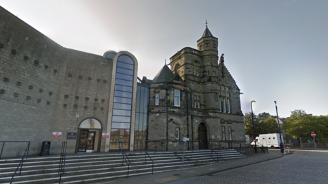 A general view of Kirkcaldy Sheriff Court. An older building made of dark stone is connected to a newer building, made of lighter stone, by several glass panels. A large brick archway is in the middle, which is also filled with glass panels.