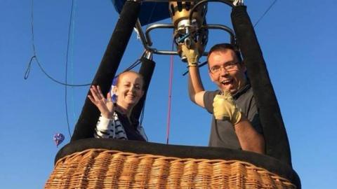 Doug and a woman in a balloon basket. He has his thumb up and his other hand on the balloon burner. His female passenger is smiling and waving at the camera
