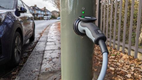 A stock image of a car being charged at a charging point, with the charger plugged into a green post in a street.