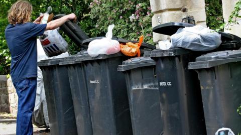 A person disposes of a bag in one of a row of black bins