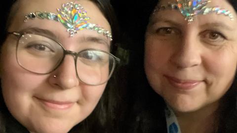 Two women looking into the camera and smiling. They are wearing stick on jewellery on their faces.