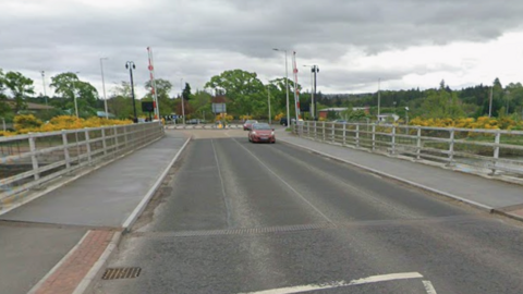 A red car is on the deck of Torvean Swing Bridge followed by another car. There are metal barriers on either side of the bridge.