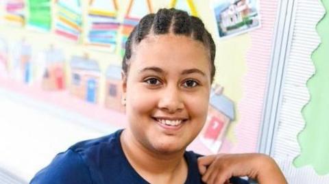 Teenage girl looks to camera. She has dark hair in cornrows and is smiling. She is wearing a blue tunic style top and is holding her left hand to her shoulder.