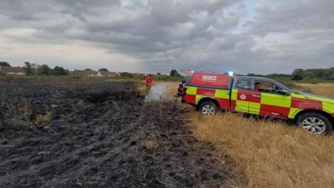An all terrain fire brigade vehicle parked by a burnt area of grassland. A firefighter is hosing down undamaged grassland adjacent to the burnt area. 