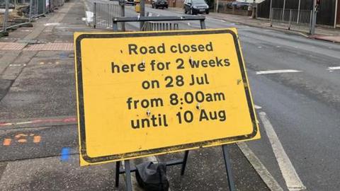A yellow highways sign which has been put by the side of the road on Mousehold Lane in Sprowston. The sign is near a set of traffic lights and pedestrian crossing. Black writing on the sign says, Road closed here for 2 weeks on 28 Jul from 8:00am until 10 Aug.