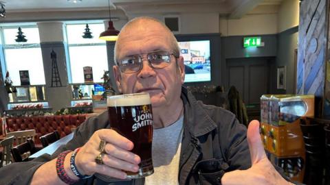 Les Sparrow holds up a point of John Smiths bitter and sticks his thump up while posing for a photo in a pub. He is wearing an open grey shirt, lighter grey jumper and glasses. 