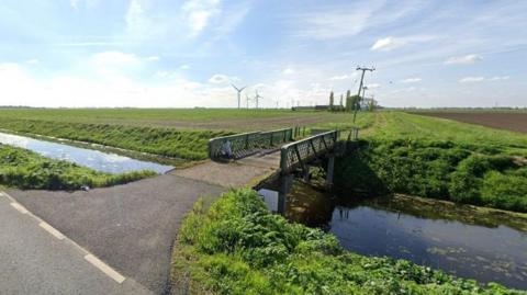 Fields span a wide landscape with a road and a bridge leading to them across a dyke, which runs parallel to a road. In the distance are wind turbines. 