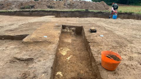 A carefully excavated trench cuts through a sandy archaeological site. Clean vertical sides reveal layers of soil and possible structural remains at the base. Tools, buckets, and tagged markers indicate an active and systematic dig. In the background, an archaeologist continues work beside mounds of removed earth.