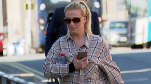 A woman with long blonde hair tied up in a ponytail, with a beige checked shirt and sunglasses, arrives at court. She has a water bottle tucked under her arm and is holding an iPhone.