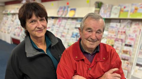 The photo shows Monika and Colin Morris standing in their shop, with shelves of greeting cards behind them.
