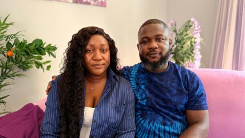 Joy Kabiri has long dark hair and is wearing a navy blue shirt with a white vest top underneath.  She is sitting next to her husband Daniel who has a beard and is wearing a navy blue t-shirt.  They are sitting on a pink sofa in their lounge.