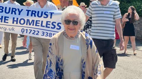 Phyll Babb, wearing a beige and blue outfit and sunglasses and with short white hair, smiling, while leading a walk. There are people behind her holding a banner for Salisbury Trust for the Homeless.