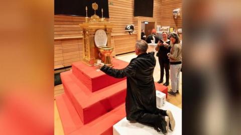 A man in a black coat and black trousers is kneeling in front of a golden toilet, which is on a raised red plinth. The toilet has golden columns and decorative detailing. The toilet is on display in an exhibition place, with two people nearby smiling and taking a photo.