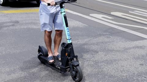 A man in sandals riding an e-scooter.