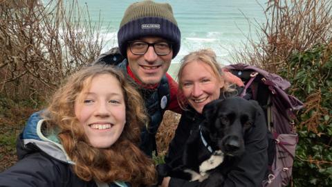 The family huddle together and smile at the camera. They are in outdoor walking gear and the sea is in the background. Tara, the mother, has the family's black and white dog on her lap.