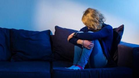 Blonde woman sits in a dark room, cradling her legs with her arms.