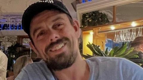 A man with a black cap, a brown beard and a light blue jumper sits in a restaurant, smiling at the camera