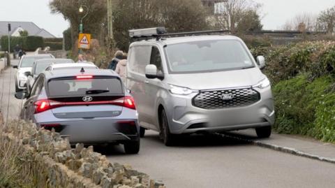 Image showing a road with part of a stone wall in the foreground. The right side of the road has a narrow pavement and a woman wearing a sling on her left arm is walking towards the camera but her head is turned looking behind at a silver van driving towards her on the pavement. Other cars are queued behind the van and one smaller car is passing in the other direction. 