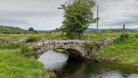 Inman's Bridge in Lyth Valley. The small bridge is made of stone and spans the narrow waterway in a single arch. Moss has grown over much of the stone work. At the centre of the arch several of the bricks have fallen slightly out of place. The bridge links two grassy fields.
