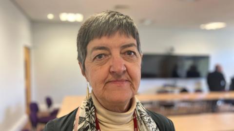Lynn Denham pictured with short grey hair while she wears a grey jacket over a cream turtle-necked jumper with a white scarf with a floral print. 