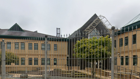 The sky is grey and there are metal silver railings in front of the court.
The court is a two storey yellowy brown building with small rectangular windows with black window panes.
There are stairs leading up to the court in a small court yard.
The pinnacle of the building is dark grey with a sharp edge on the arch. 