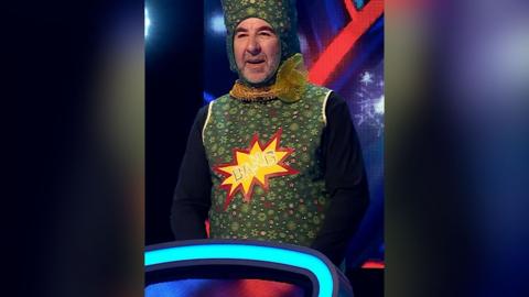 A man stands behind a podium wearing a giant green cracker outfit