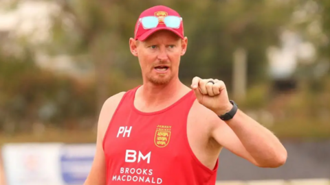 Paul Hutchinson wearing a red cap and red vest with the Jersey cricket crest on