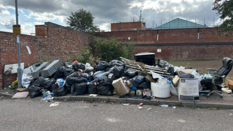 A picture showing black bin bags, a chair, cardboard boxes and a metal frame piled on a concrete curb.