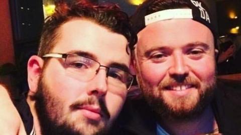 Two young men with their arms around each others shoulders smile at the camera. The one on the left is wearing glasses and the one on the right has a cap on backwards