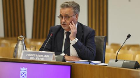 Prof Iain Gillespie answers questions of MSPs, sits at committee room table