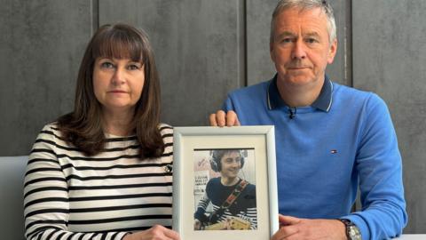 Niamh Dolan, wearing a black and white striped top, and her husband Peter Dolan, wearing a blue jumper over a dark blue polo shirt, both sit looking towards the camera, holding a photo of their late son Enda, who is pictured playing guitar and smiling. 