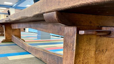 Close-up of a long, heavy wooden table with thick legs and a horizontal support beam, placed on a carpet with multicoloured stripes. The wood shows signs of age and wear, and the year ‘1937’ is carved into one of the legs.