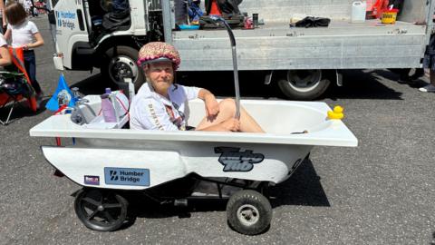 A man wearing a white T-shirt, shorts and a colourful helmet sat inside a kart made out of a bath. 