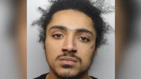 A police mugshot of a man with black hair which is sticking up in places. He has short stubble and is wearing a black T-shirt.