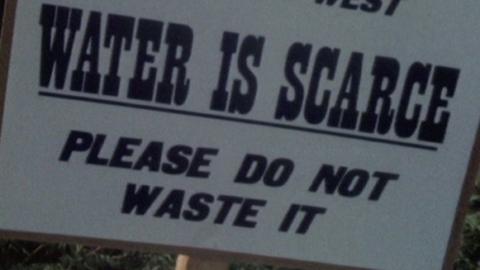 A white sign is attached to a wooden post. In bold letters it reads: "Visitors Welcome to the South West, Water is scarce. Please do not waste it."