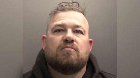 Police mugshot of Danny McNaughton, who has his head raised, has long brown stubble and moustache and messy short brown hair on top of his head with short sides.