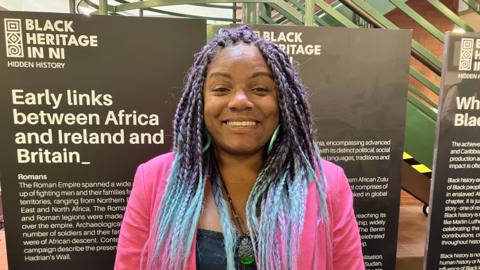 Woman has long and colourful blue and purple hair. 
She is wearing a bright pink jacket and green hoop earring and is smiling. 
She is standing in front of placards for the Black Heritage NI project which are positioned in front of a green staircase. 