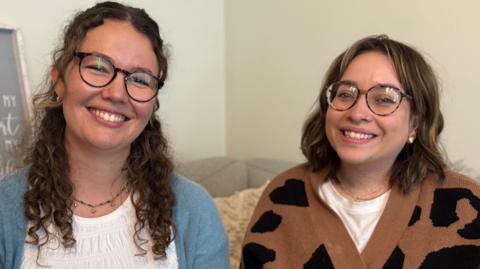 Two young women, both wearing glasses, smile at the camera. The lady on the left has long curly brown hair and is wearing a white t-shirt with a blue cardigan. The lady on the right has shoulder length brown hair and is wearing a white t-shirt with a brown and black animal print cardigan over the top.