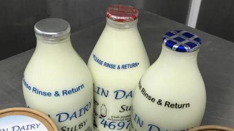 Three milk bottles full with white milk, with foil tops. One is silver, another is red, the third is blue.