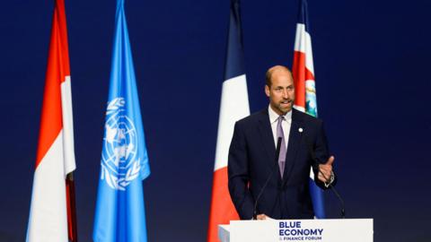 prince william at a podium in front of flags