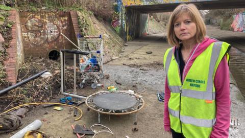 Professor Sarah Gabbott from the University of Leicester is in a hi-viz top and standing beside the Willow Brook waterway in Leicester. Alongside the channel of water, there is an array of junk and rubbish, including an old shopping trolly and a small child's trampoline. 